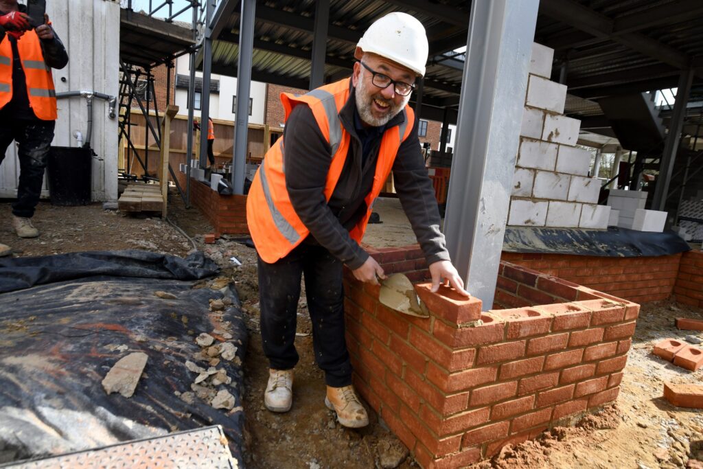 Image by Vikki Lince - a man in a hard hat and high vis jacket has laid a brick for the new Grove Cottage building